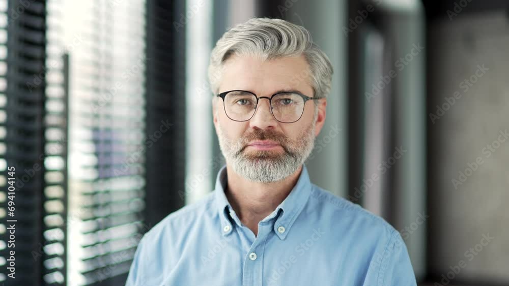 Portrait of serious mature gray haired bearded businessman standing at workplace in business office. Confident senior handsome teacher in glasses looking at camera. Head shot of professor. Close up Portrait of serious mature gray haired bearded businessman standing at workplace in business office. Confident senior handsome teacher in glasses looking at camera. Head shot of professor. Close up