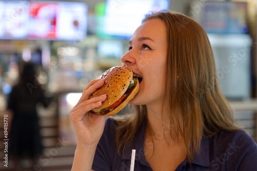 Portrait of young happy woman or beautiful teenager girl eating fast junk food  tasty burger and drink soda in a restaurant or cafe  enjoy meal. 