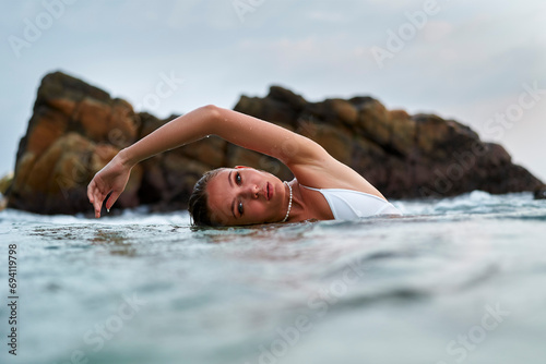 Woman luxuriates in tranquil sea, swims near rocks in lagoon. Female enjoying serene waters, leisure swim, nature beauty. Model showcases waterproof makeup, water-resistant attire in ocean setting. photo