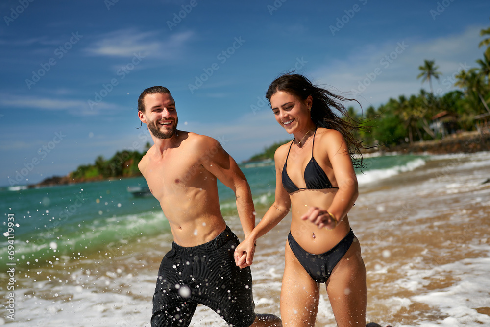 Joyful couple runs on tropical beach, splashing water. Shirtless man with athletic body leads, slender woman in bikini follows. Fun, fitness on seaside. Active, attractive pair enjoys summertime. Joyful couple runs on tropical beach, splashing water. Shirtless man with athletic body leads, slender woman in bikini follows. Fun, fitness on seaside. Active, attractive pair enjoys summertime.