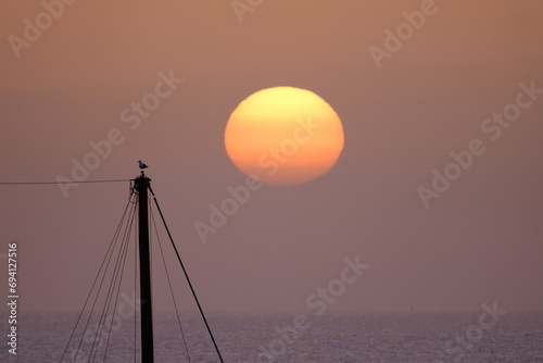 A beautiful bright sunset on the ocean, a huge sun setting behind the horizon against the background of the silhouette of a ship’s mast and a bird on it. Fuerteventura, Canary Islands.