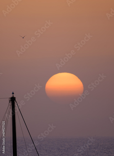 A beautiful bright sunset on the ocean, a huge sun setting behind the horizon against the background of the silhouette of a ship’s mast and a bird on it. Fuerteventura, Canary Islands.