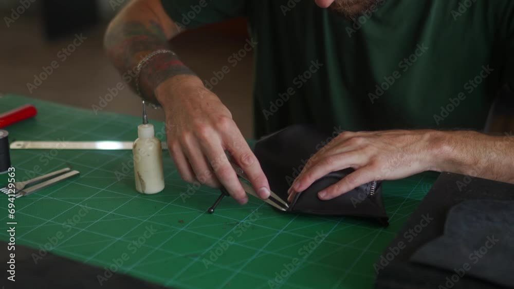 Close up shot of professional leather craftsman's hands at work table making cut on black handbag with wing divider. Man in khaki T-shirt sewing purse in workshop. Close up shot of professional leather craftsman's hands at work table making cut on black handbag with wing divider. Man in khaki T-shirt sewing purse in workshop.