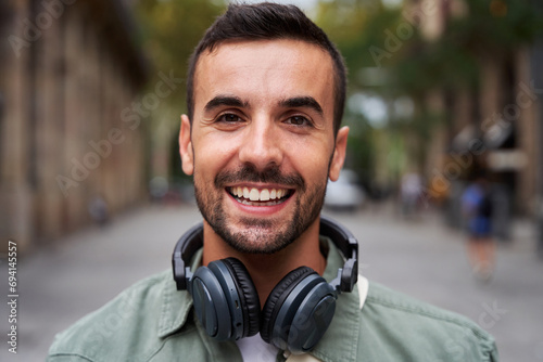 Portrait of a young handsome man smiling and looking at camera. Happy male wearing headphones in street on the city. Close up of a cheerful millennial caucasian guy outdoors. photo