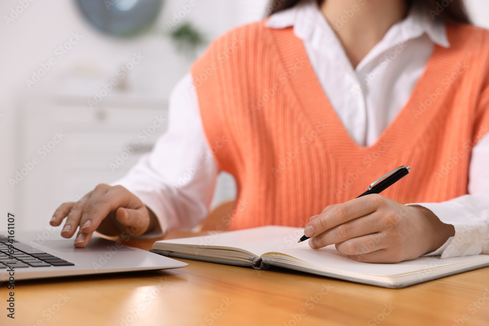 Young woman writing in notebook while working on laptop at wooden table, closeup Young woman writing in notebook while working on laptop at wooden table, closeup