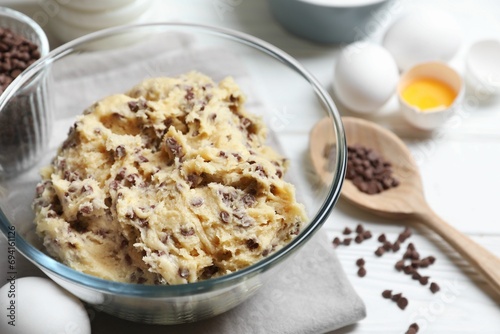 Chocolate chip cookie dough in bowl and ingredients on white wooden table, closeup