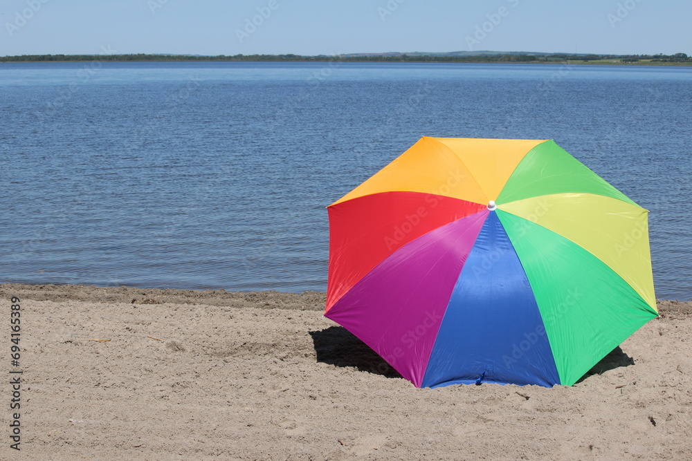 Summer time beach and vacation vibes created by a vibrant rainbow colored umbrella lying on the sandy beach at the lake intentionally offset for copy space.