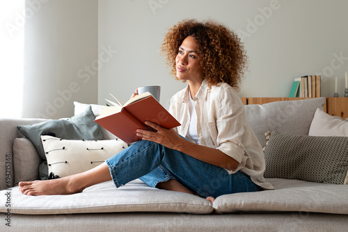 Pensive relaxed African american woman reading a book at home, drinking coffee sitting on the couch. Copy space. photo