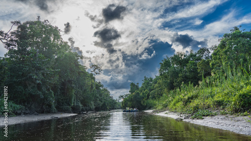The river flows through a protected rainforest. A boat with tourists moored to the shore. Thickets of lush tropical vegetation on the banks of the river. The blue sky, clouds are reflected