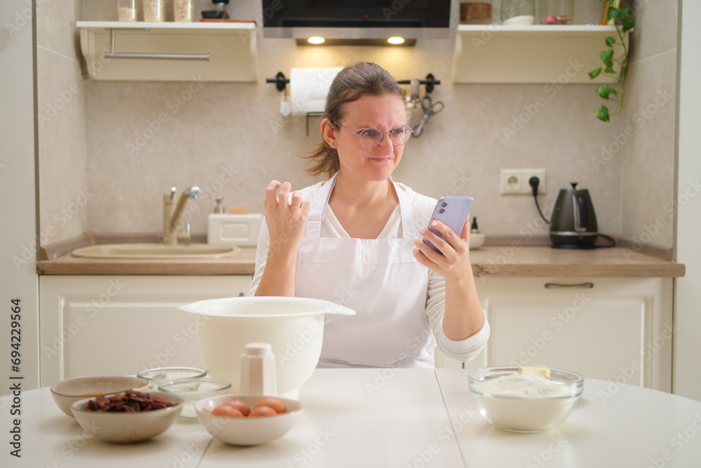 Angry woman making dough in the kitchen at home. She is using a smartphone. Process of cooking pecan pie in home kitchen for American Thanksgiving Day.