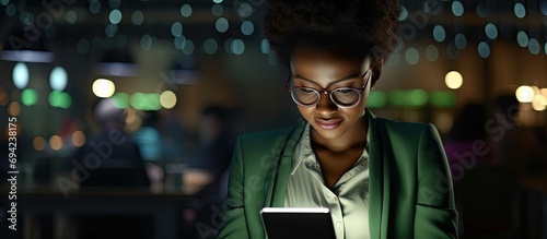 A Nigerian woman in a dark office is using a tablet for business-related tasks at night, such as meeting deadlines and conducting online research, as well as engaging in social media and internet