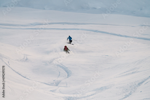 Two skiers complete a joint descent down a snowy slope, one of them jumps over the springboard