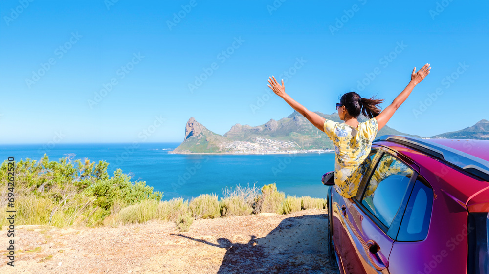 woman outside a car window with hands up, a car at Chapman's Peak Drive in Cape Town South Africa looking out over the ocean. women on a road trip at the garden route South Africa with a renal car - obrazy, fototapety, plakaty 