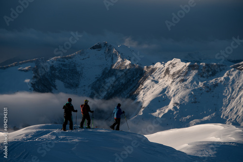 Three male skiers stand on a snow-covered mountain peak and look at another mountain peak
