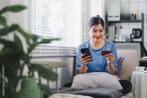 Beautiful Asian young woman using smartphone for online shopping and making a payment with credit card while sitting on the sofa at home. internet banking applications and e-commerce concept.