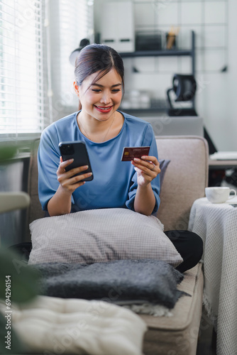 Beautiful Asian young woman using smartphone for online shopping and making a payment with credit card while sitting on the sofa at home. internet banking applications and e-commerce concept. © kenchiro168