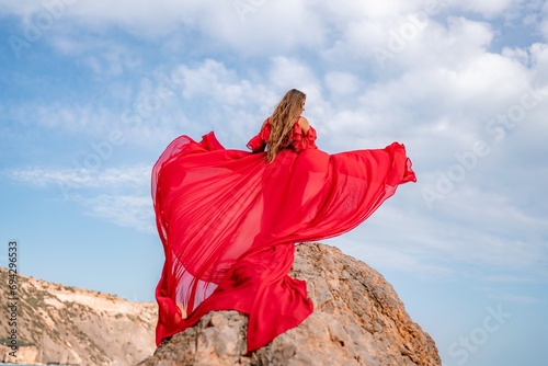 woman sky red dress. Woman with long hair on a sunny seashore in a red flowing dress, back view, silk fabric waving in the wind. Against the backdrop of the blue sky and mountains on the seashore.