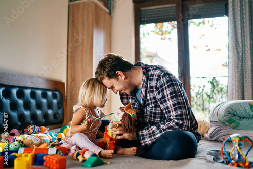 Dad and little girl are sitting on the bed touching their foreheads playing toys