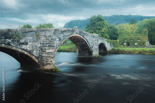 Old Roman inspired bridge Pont Fawr across the River Conwy towards a 15th century ivy covered stone home in the Welsh village of Llawnrst, North Wales photo