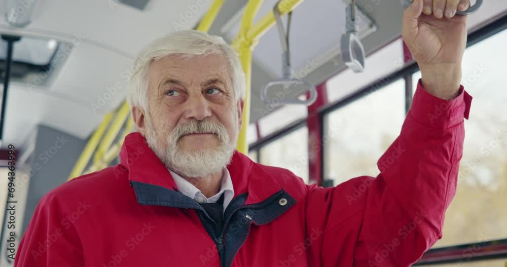 Front view of old man standing on bus, looking around. Male with grey hair and beard, wearing red jacket, holding bus handle, smiling. Concept of modern urban routine and life.