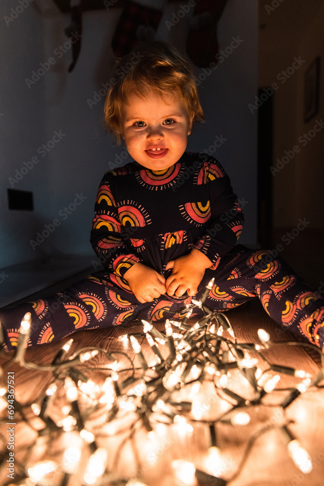 Two year old baby girl playing with Christmas lights on the floor. Smiling in beautifull dress. Dark background. Being curious. Blonde hair, blue eyes. Two year old baby girl playing with Christmas lights on the floor. Smiling in beautifull dress. Dark background. Being curious. Blonde hair, blue eyes.