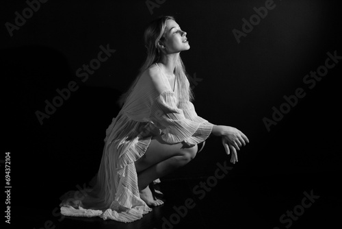 Low key black and white portrait of young pretty woman in vintage white dress sitting on the floor