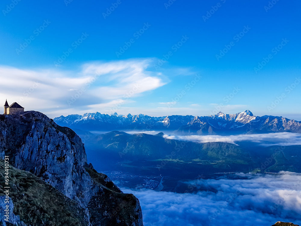 Scenic sunrise view of chapel Maria am Stein on top of mountain peak Dobratsch, Villacher Alps, Austria, Europe. Looking at Julian and Karawanks mountain range. Golden morning hour tranquil atmosphere