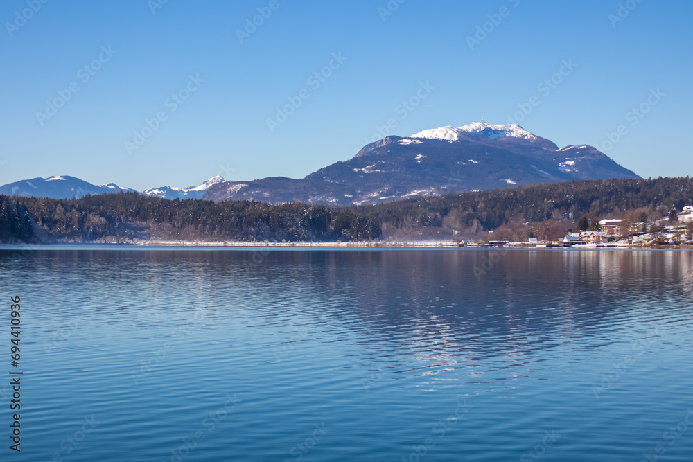 Naklejka premium Panoramic view on lake Faakersee in Austrian Alps, Carinthia, Austria. Lake is surrounded by high snow capped mountains. Calm water surface with reflections of landscape. Looking at Dobratsch peak