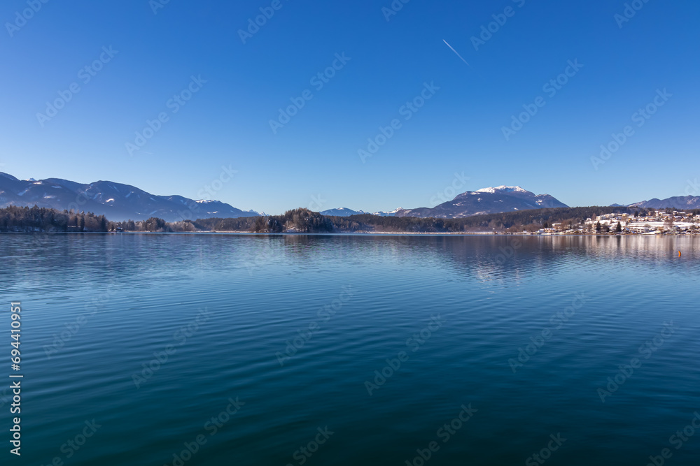 Panoramic view on lake Faakersee in Austrian Alps, Carinthia, Austria. Lake is surrounded by high snow capped mountains. Calm water surface with reflections of landscape. Looking at Dobratsch peak