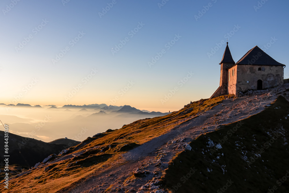 Scenic sunrise view of chapel Maria am Stein on top of mountain peak Dobratsch, Villacher Alps, Austria, Europe. Looking at Julian and Karawanks mountain range. Golden morning hour tranquil atmosphere