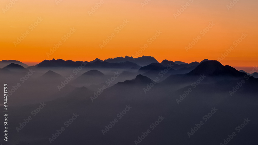 Panoramic sunrise view from Dobratsch on Julian Alps and Karawanks in Austria, Europe. Silhouette of endless mountain ranges with orange and pink sky. Jagged sharp peaks and valleys. Cottage on hill