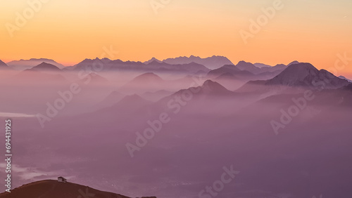 Panoramic sunrise view from Dobratsch on Julian Alps and Karawanks in Austria  Europe. Silhouette of endless mountain ranges with orange and pink sky. Jagged sharp peaks and valleys. Cottage on hill