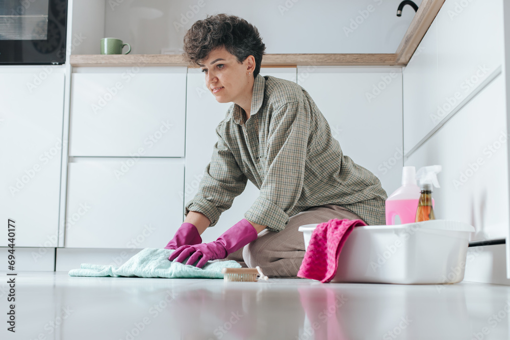 A Caucasian woman with a short haircut washes the floor in lilac gloves in a light kitchen. House cleaning products are nearby. A Caucasian woman with a short haircut washes the floor in lilac gloves in a light kitchen. House cleaning products are nearby.
