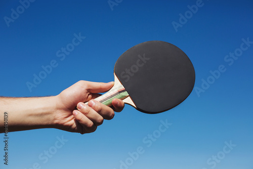 Hand holding black ping pong racket on blue sky background, closeup. Ping pong paddle. Playing ping pong.  photo