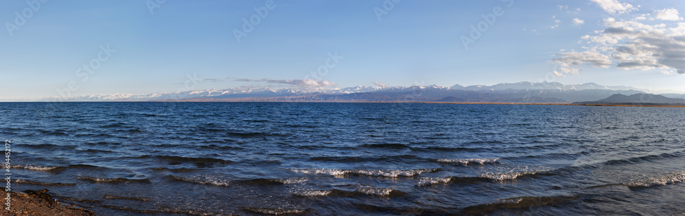 Blue calm water in Issyk-Kul lake with mountains on background at summer day - wide panorama.