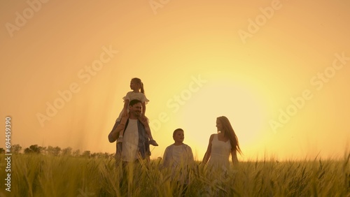 Happy family with child go through wheat field holding hands. Slow motion. Mom, dad and children are walking together. Active mother, father and little daughter play, enjoy nature outdoors, dream. Kid