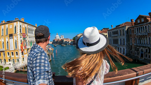 Tourist couple standing on top of famous Rialto bridge overlooking the Canal Grande in Venice, Veneto, Northern Italy, Europe. Female model is wearing black dress. Romantic luxury summer vacation