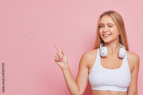 Pretty blondie with headphones on her neck points finger aside winking and smiling over background, good choice concept, copy space