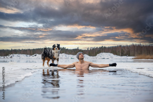 Young man taking an ice bath with his dog suring sunset. Swimming in a frozen lake in winter. Healthy cold swim.