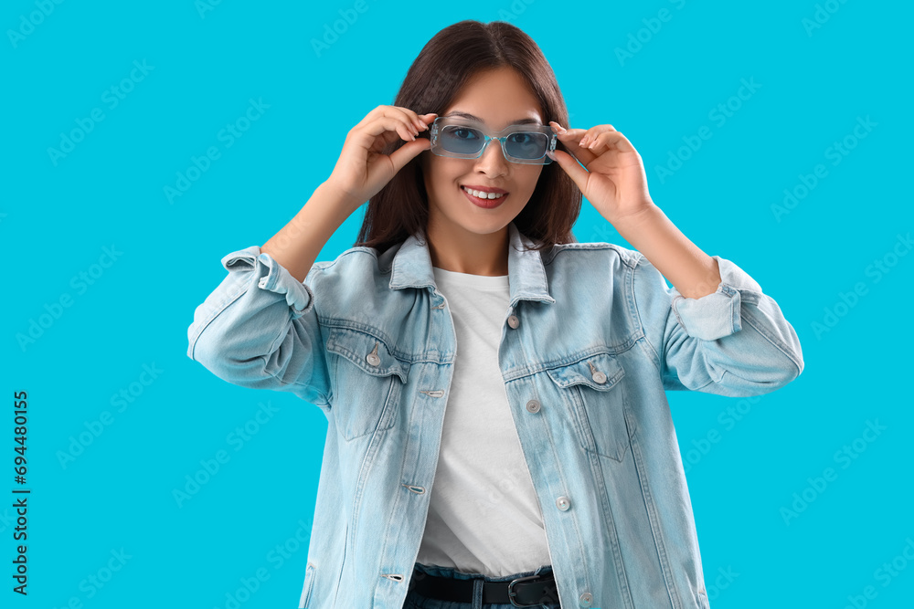 Stylish young Asian woman in sunglasses and denim jacket on blue background