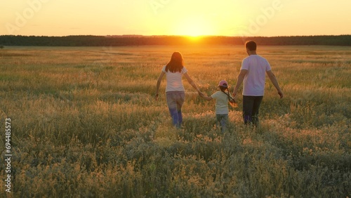 Family walks on green grass in meadow. Happy family  child  are walking in summer field  holding hands. Mom dad daughter walking together on nature. Parents  children are walking in park at sunset.