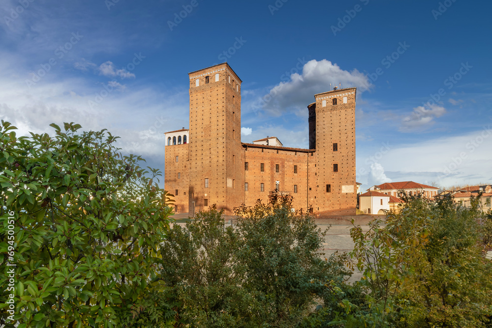 Fossano, Italy - Castle of the Princes of Acaja (14th century) in Castle Square over blue sky with white clouds, green hedge in foreground