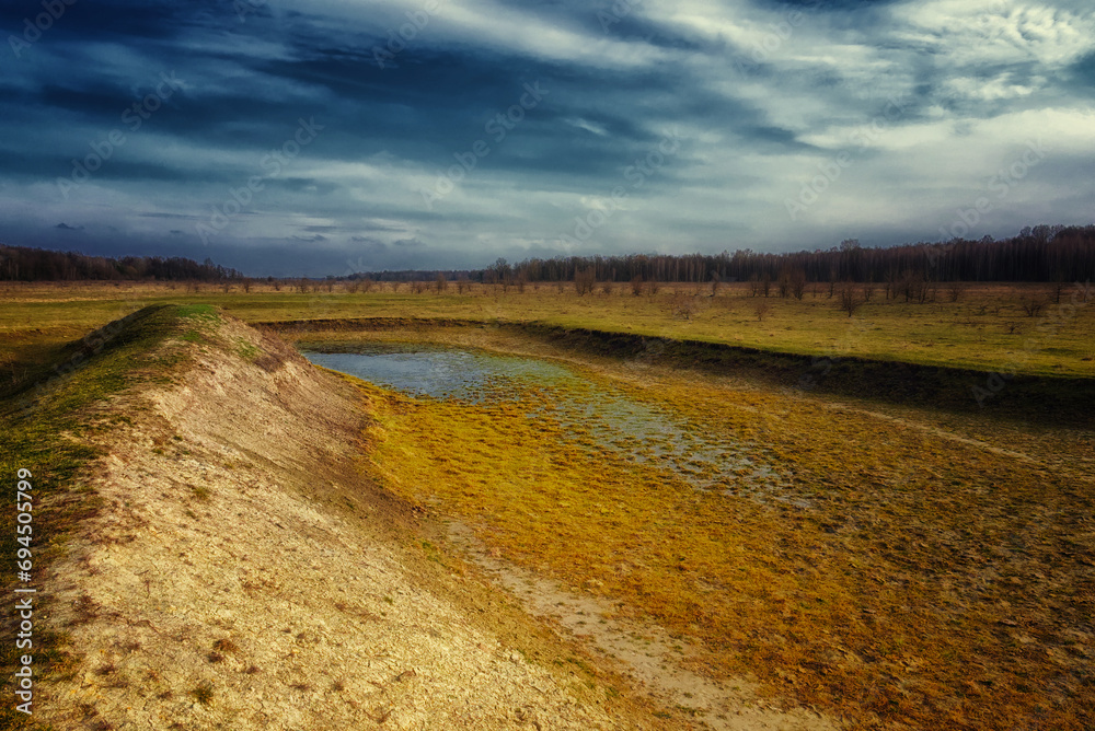 The image showcases a field with a small body of water, a dirt path meandering towards the horizon. The exposed bottom of a dry pond. A shallow water body.