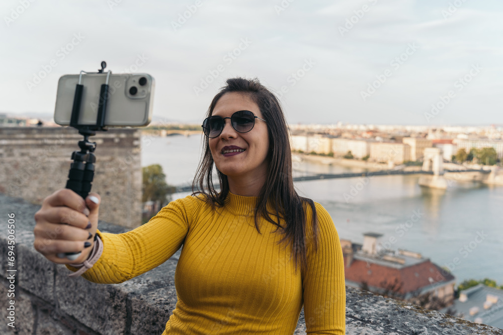 Fototapeta premium Young content creator filming herself with her cell phone on a tripod while talking to her followers. Girl filming herself with her cell phone to show the city of Budapest.