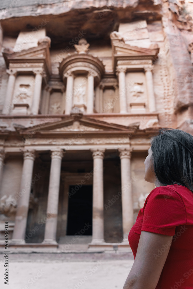Young European tourist looking at the Treasury of Petra, Jordan. Brunette girl in red dress enjoying the view of the façade and the ruins of the ancient city carved in stone.
