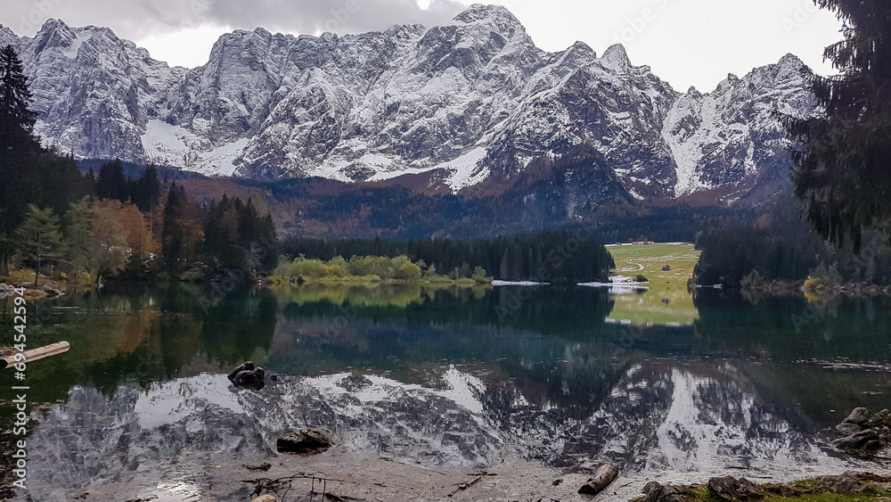 Fototapeta premium Panoramic view of Fusine Lake (Laghi di Fusine) and snow capped Julian mountain range in Tarvisio, Friuli-Venezia Giulia, Italy, Europe. Water reflection in green alpine lake. Mountain peak Mangart