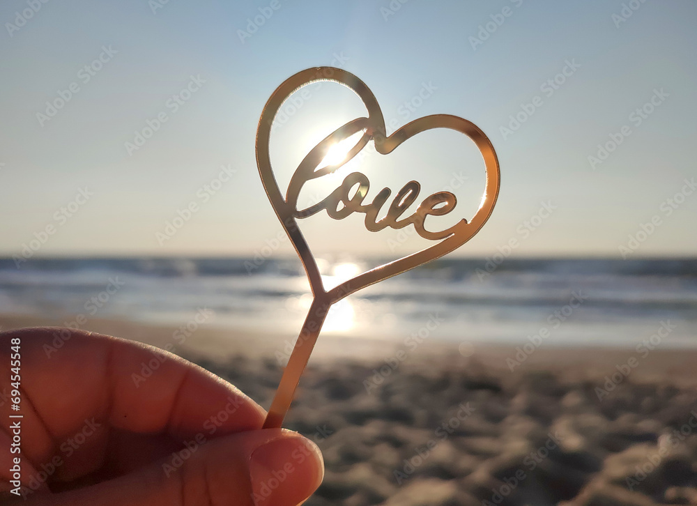 Fototapeta premium Person holding in fingers hand stick in shape golden heart and word Love on background sea and sea waves in seashore on sunny summer day close-up. Concept love romance amour St Valentines Day