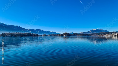 Snow covered wooden pier on lake Faak in Carinthia, Austria, Europe. Surrounded by high snow capped Austrian Alps mountains. Calm water surface with reflections of landscape. Looking at Dobratsch peak