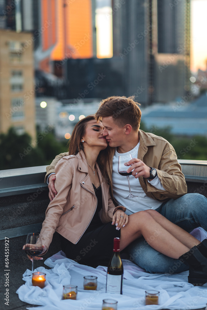 Fototapeta premium Romantic beautiful surprise on Saint Valentine's Day for a girlfriend. Loving young couple having dinner on a rooftop on sunset evening with candles, wine. Skyscrapers on background, urban cityscape