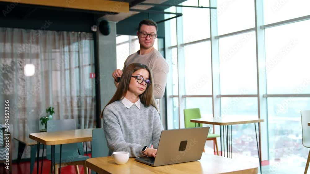 Long-haired female working at her laptop in the office. Man stirring sugar in the cup comes up behind peeping at colleagues computer. Long-haired female working at her laptop in the office. Man stirring sugar in the cup comes up behind peeping at colleagues computer.